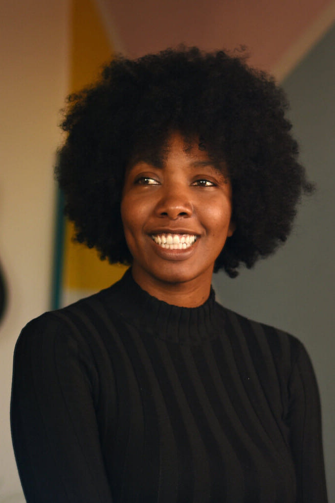 photographie d'Amandine Gay, jeune femme Noire avec une coupe afro, vêtue de noir, souriant